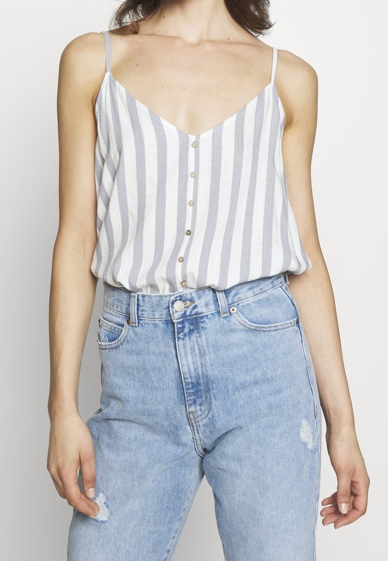 Randig ärmlös topp med v-ringning; ljusgrå och vita färger; har små knappar fram och en avslappnad passform. Bärs med blå jeans.