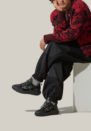 Sneakers neri con texture e accenti grigi, indossati con calzini grigi e pantaloni neri larghi. Maglione a righe rosse e nere in vista. Sfondo semplice.