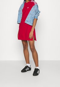 Červené šaty s bočními rozparky, doplněné o modrou džínovou bundu s lehkým praním a třemi bílými pruhy, k tomu černé tenisky s bílými akcenty.