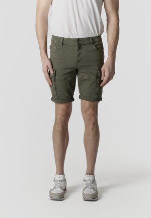 Hombre vistiendo pantalones cortos cargo verde oliva, zapatillas blancas y una camiseta blanca, de pie contra un fondo claro y liso.