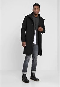 Homme portant un manteau noir, une chemise noire à motifs, un jean gris roulé et des bottes noires, se tenant contre un fond blanc.