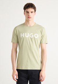 Šviesiai žalias medvilninis marškinėlis su dideliu baltu "HUGO" logotipu priekyje. Trumpomis rankovėmis ir apvaliu iškirpimu, glotnus paviršius.