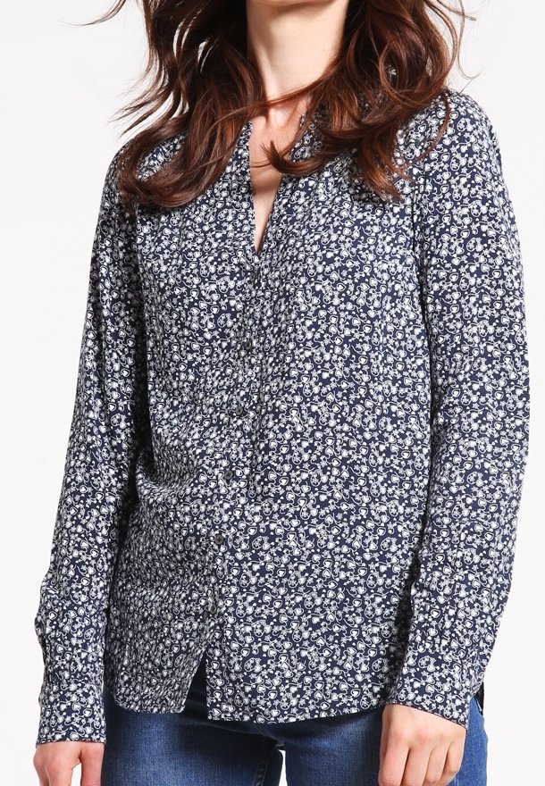 Femme portant un chemisier bleu marine à manches longues avec un petit motif floral blanc et un jean bleu, debout devant un fond uni.