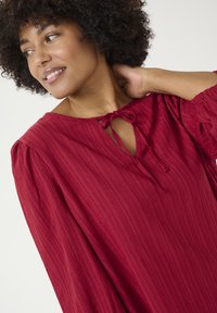 Red textured blouse featuring a keyhole opening with a tied detail at the neckline and puffy sleeves. Fabric has a subtle striped pattern.
