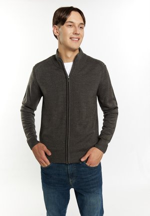 Sudadera gris con cremallera y cuello alto, con puños acanalados, hecha de un tejido de punto texturizado. Lleva puesta sobre una camiseta blanca y jeans azules.