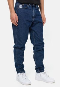 Hombre vestido con pantalones de mezclilla azul oscuro y zapatillas blancas, de pie con las manos relajadas a los lados sobre un fondo blanco.