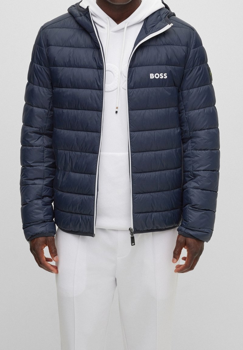 Homme portant une veste matelassée bleu marine avec le logo "BOSS" par-dessus un sweat à capuche blanc et un pantalon blanc, debout devant un fond uni.