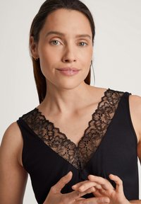 Canotta nera con scollatura a V in pizzo nero trasparente. Il pizzo presenta motivi floreali e bordi ondulati, mettendo in risalto il design della scollatura.