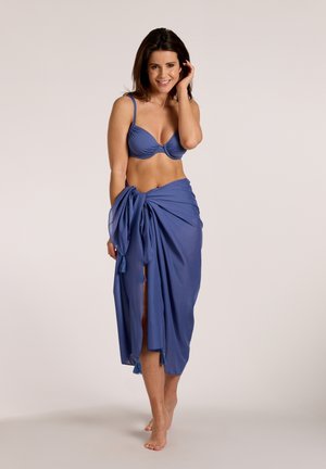 Woman smiling, wearing a blue bikini top and matching sarong tied at the waist, standing barefoot against a plain background.