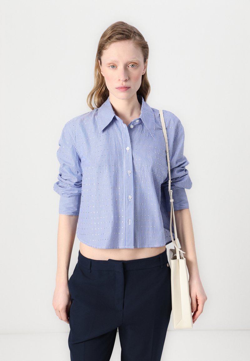 Camicia cropped a righe blu e bianche con bottoni frontali, maniche lunghe arrotolate, abbinata a pantaloni blu navy e una borsa color crema.