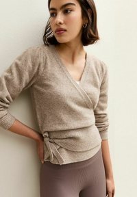 Frau in beiger Wickelstrickjacke mit seitlichem Bindeband und braunen gerippten Leggings, die an einer schlichten hellen Wand steht und zur Seite schaut.