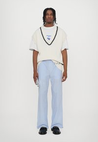 Gilet bianco a maglia con rifiniture nere sopra una maglietta bianca, abbinato a pantaloni a gamba larga a righe blu e bianche e scarpe nere. Tiene un oggetto piccolo.