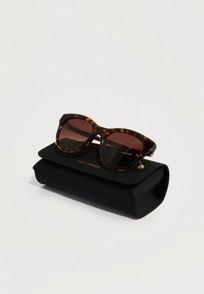 Slnečné okuliare so želvovým vzorom s tmavými šošovkami na čiernom mäkkom púzdre, s detailom zlatého loga a zaobleným dizajnom rámu.