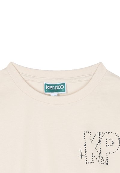 T-shirt couleur crème avec un col rond, présentant un logo "KP" noir à pois et des embellissements, fabriqué en coton doux.