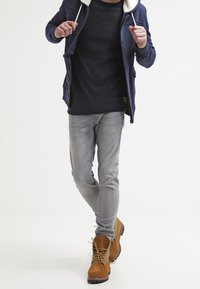 Chaqueta azul marino con cremallera, cuello de borrego blanco, camisa negra de manga larga, jeans ajustados gris claro y botas de trabajo color canela con suelas negras.