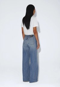Femme aux longs cheveux noirs debout, portant un haut blanc à manches courtes et un jean bleu taille haute à jambes larges, face à un mur blanc.