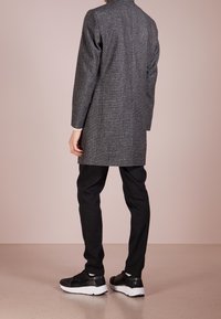 Grå houndstooth frakke med høj krave, parret med sorte bukser og sorte sneakers med hvide såler, set fra ryggen.