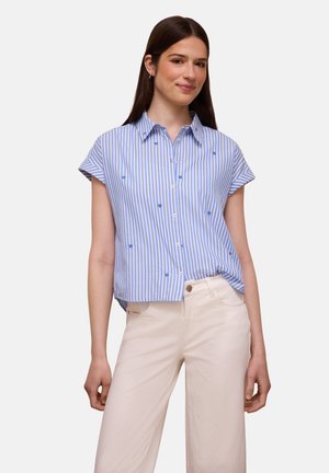 Młoda kobieta w niebiesko-białej pionowo paskowanej koszuli na krótkie rękawy z małymi wzorami oraz jasnobeżowych spodniach.