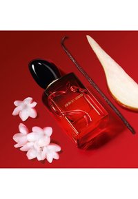 Bottiglia di profumo rossa con tappo nero, caratterizzata da un design floreale. Accompagnata da petali rosa, un baccello di vaniglia e una fetta di pera.