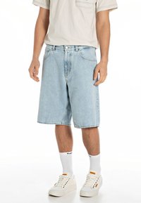 Pantalones cortos de mezclilla de color azul claro con un corte relajado, que presentan bolsillos frontales y un solo botón de cierre. Combinados con zapatillas blancas y calcetines cortos.