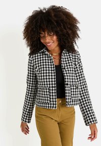 Veste à motif pied-de-poule noir et blanc avec une coupe courte, des fermetures à boutons et un col, accompagnée d'un haut noir et d'un pantalon beige.