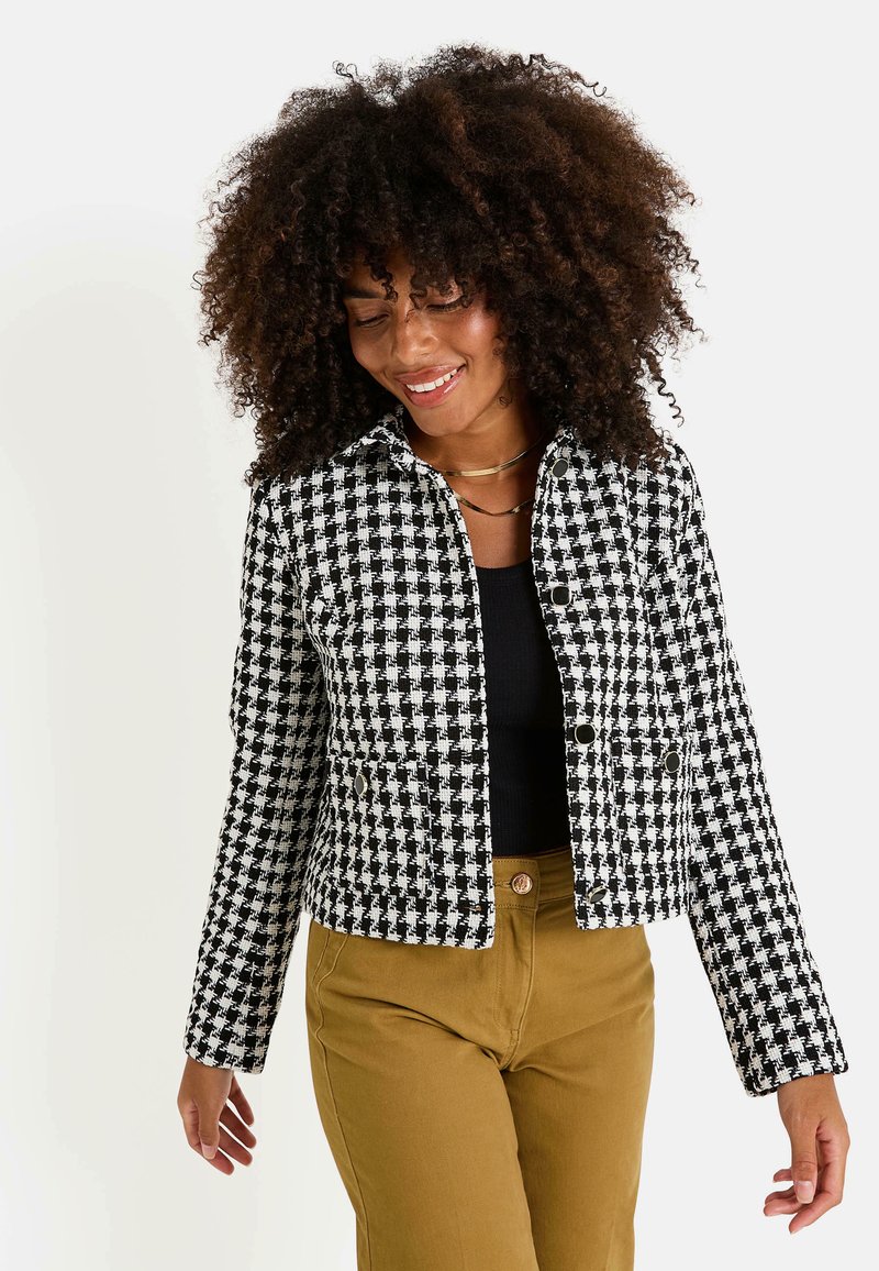 Veste à motif pied-de-poule noir et blanc avec une coupe courte, des fermetures à boutons et un col, accompagnée d'un haut noir et d'un pantalon beige.