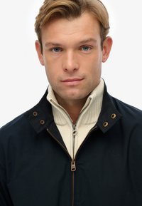 Chaqueta navy con herrajes dorados, un cuello alto y un suéter crema con cremallera debajo. Tela texturizada y detalles de costura visibles.