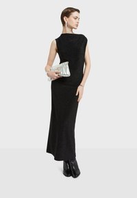 Sort, ærmeløs kjole i gulvlængde med en tætsiddende pasform og diskret glimmer. Modellen holder en sølvfarvet clutch og har sorte ankelstøvler på.