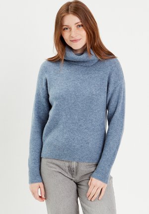 Pull en tricot doux, bleu clair, avec un col roulé haut, des manches longues et des bords côtelés aux poignets et à l'ourlet. Porté avec un pantalon gris.