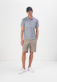 Grau karierter Poloshirt mit einem einfarbigen grauen Einsatz; kombiniert mit beigen Shorts und weißen Sneakers. Schwarze Mütze auf dem Kopf. Einfacher, lässiger Look.