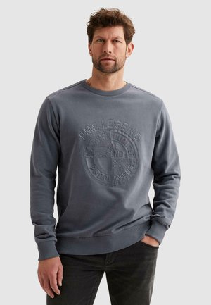 Mann mit Bart, der einen dunkelgrauen Pullover mit eingeprägtem "PME Legend"-Logo trägt und vor einem schlichten hellen Hintergrund steht.