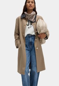 Cappotto a quadri marrone con colletto nero, sciarpa crema e felpa bianca. Jeans a vita alta blu e borsa chiara con plissettature.