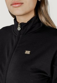 Giacca nera con zip in tessuto morbido e colletto alto. Presenta una zip in metallo argentato e un piccolo logo metallico sul petto.