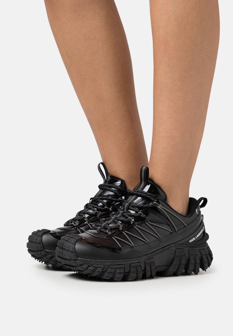 Personne portant des baskets noires à semelles épaisses avec des détails en cuir verni et des lacets, debout sur un fond clair uni.