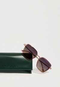 Stačiakampių formų „Lacoste“ akiniai nuo saulės su rožinės spalvos lęšiais ir auksiniais akcentais, padėti šalia tamsiai žalių odinių dėklų.