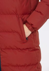 Rød isoleret jakke med en glat tekstur, fyldig design, elastisk manschetter og sidelomme med et blåt accent på lynlås trækkeren.