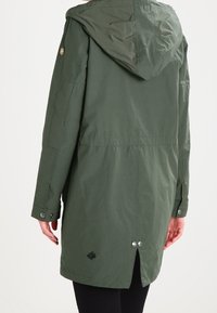 Mörkgrön huva jacka, gjord av slätt tyg, har en bakventil med tryckknappar och en emblemdetalj på nedre vänstra sidan.