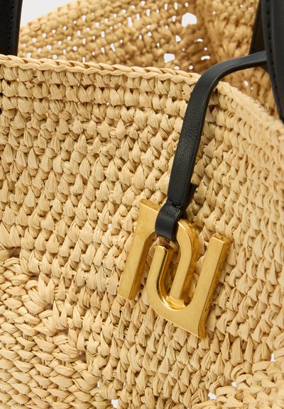Πλεκτή τσάντα από άχυρο με μαύρη δερμάτινη λαβή και προσαρτημένη χρυσή γεωμετρική κρεμαστή λεπτομέρεια.