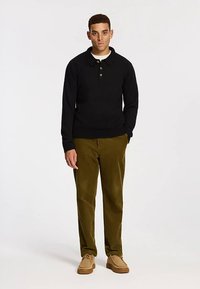 Zwarte polo met lange mouwen en drie knopen, gecombineerd met olijfgroene corduroy broek en bruine schoenen. Model staat in een neutrale houding.