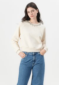Felpa crema con scollo decorato, polsini e vita a costine. Abbinata a jeans in denim a taglio rilassato di colore azzurro.