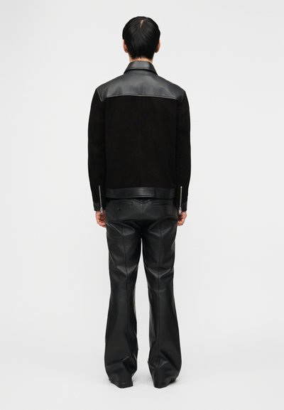 Άτομο που στέκεται πλάτη, φορώντας μαύρο δερμάτιν ο παντελόνι και μαύρο μπουφάν με δερμάτινες λεπτομέρειες στους ώμους και το τελείωμα, πάνω σε λευκό φόντο.