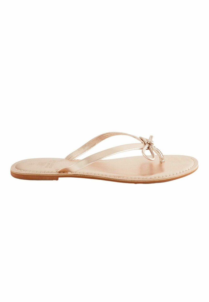 Nude-värinen flip-flop, jossa on kaksi kapeaa remmiä, jotka risteävät jalan päällä, litteä nahkapohja ja koristeellinen solmi yksityiskohta varpaassa.