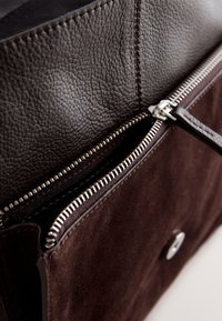 Interiorul unei genți de mână din piele maro și piele întoars ă, având o închidere cu fermoar argintiu, suprafață texturată și un detaliu metalic cu buton.