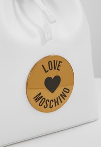 Vitt läderväskan med ett guld cirkulärt emblem som har ett svart hjärta och orden "LOVE MOSCHINO" i svarta bokstäver.