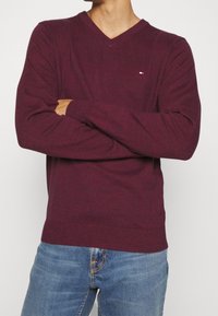 Burgundowy sweter w serek wykonany z miękkiego dzianiny, cechujący się dopasowanym krojem i małym logo na klatce piersiowej. W połączeniu z niebieskimi dżinsami.