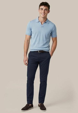 Jonge man die lacht, draagt een lichtblauw polo met korte mouwen, marineblauwe broek, een donkere riem en bruine instapschoenen, staat met één hand in de zak.