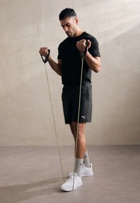 En svart kortermet skjorte og grå shorts båret av en mann som bruker gule motstandsbånd. Han har på seg hvite sko og lysegrå sokker.