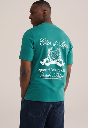 Mann trägt ein türkisfarbenes T-Shirt mit weißem Schriftzug "Côte d'Azur Sports & Leisure Club" und Emblem auf dem Rücken, kombiniert mit dunkelblauer Jeans.