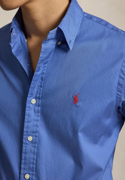 Homme portant une chemise bleue à boutons avec un logo rouge brodé d'un joueur de polo sur le côté gauche de la poitrine.