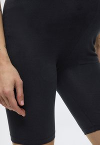 Prim-plan cu o persoană purtând pantaloni scurți negri mulați, arătând mâna stângă odihnindu-se lângă coapsă și un tatuaj mic pe antebraț.